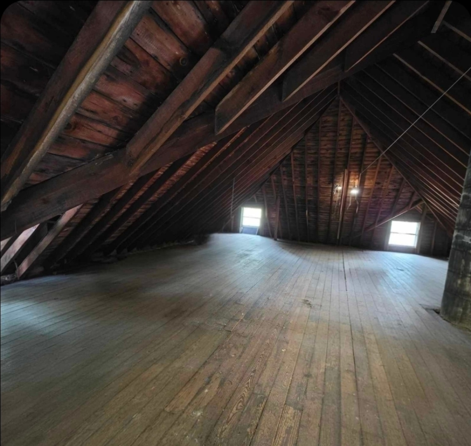 An unfinished Victorian attic with exposed wooden beams and flooring, filled with natural light from two windows. The bare wooden structure shows the bones of the 1914 house that will become a creative studio space.