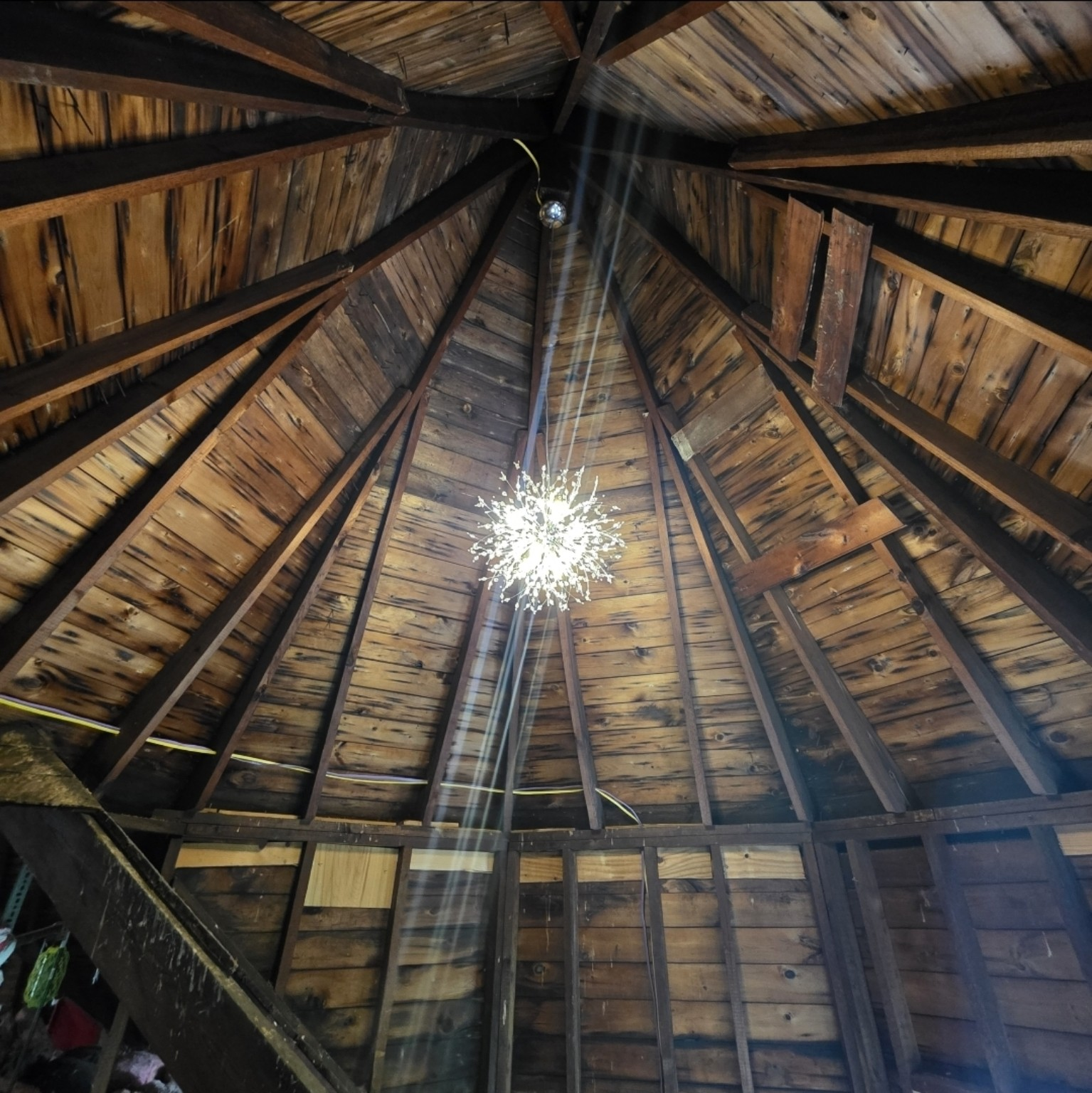 Looking up into the unfinished Victorian turret ceiling with original 1914 wooden beams radiating outward, and a sparkling chandelier already hanging in the center.